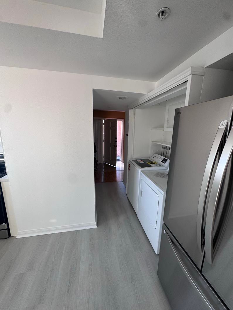 Narrow kitchen with stainless steel refrigerator, white cabinets, and gray flooring leading to a hallway.