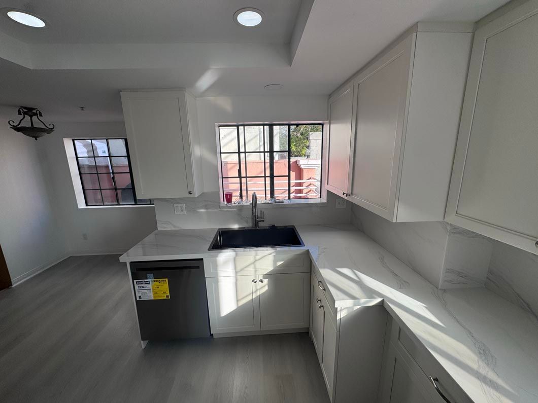 Bright white kitchen with island, sink, cabinets, and large windows letting in sunlight.