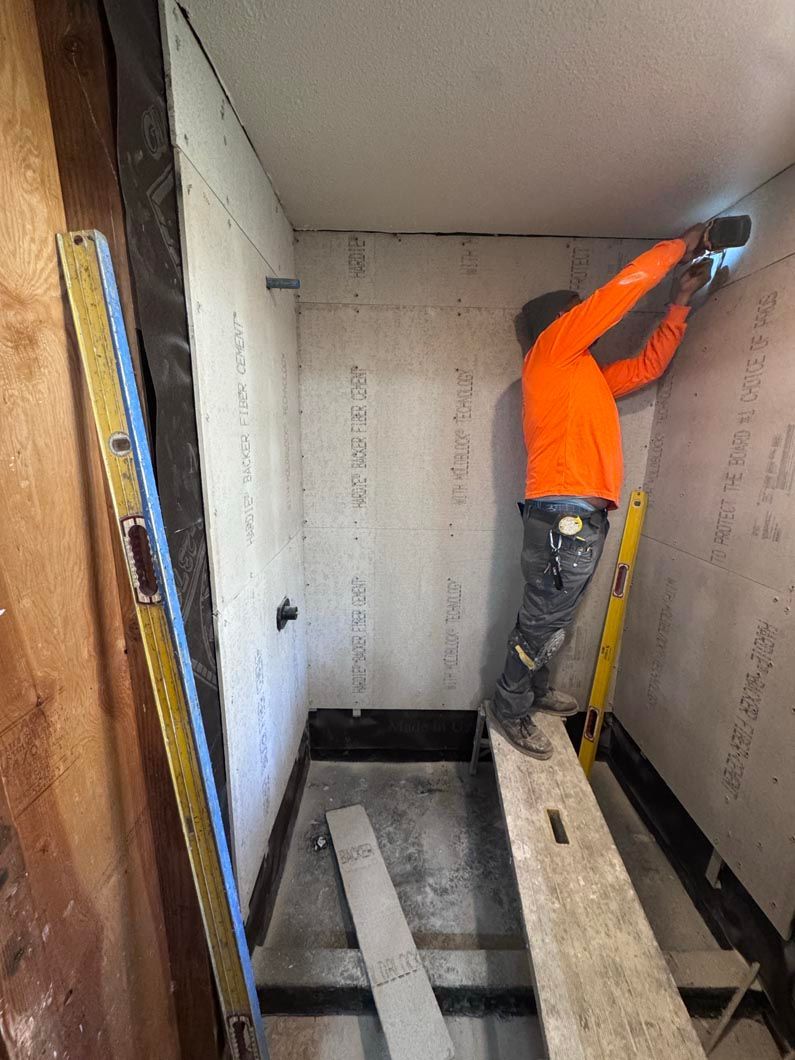 Worker in orange jacket installing drywall on a small interior room wall using a ladder.