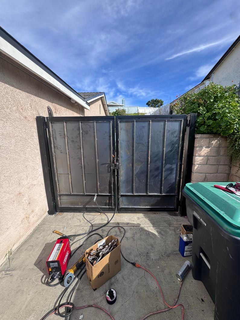 Narrow outdoor alley with a closed black metal gate, scattered tools, and trash bins on the right.