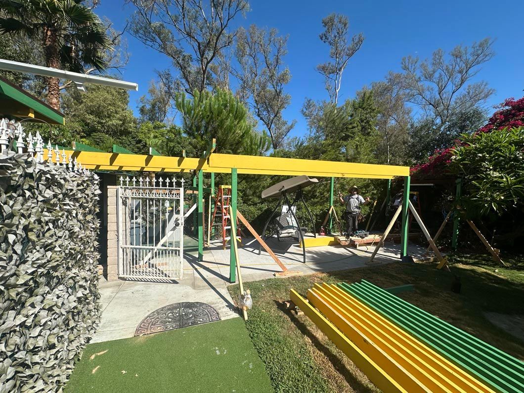 Outdoor yard with a yellow canopy over a gated patio and green-yellow benches, surrounded by trees and flowers