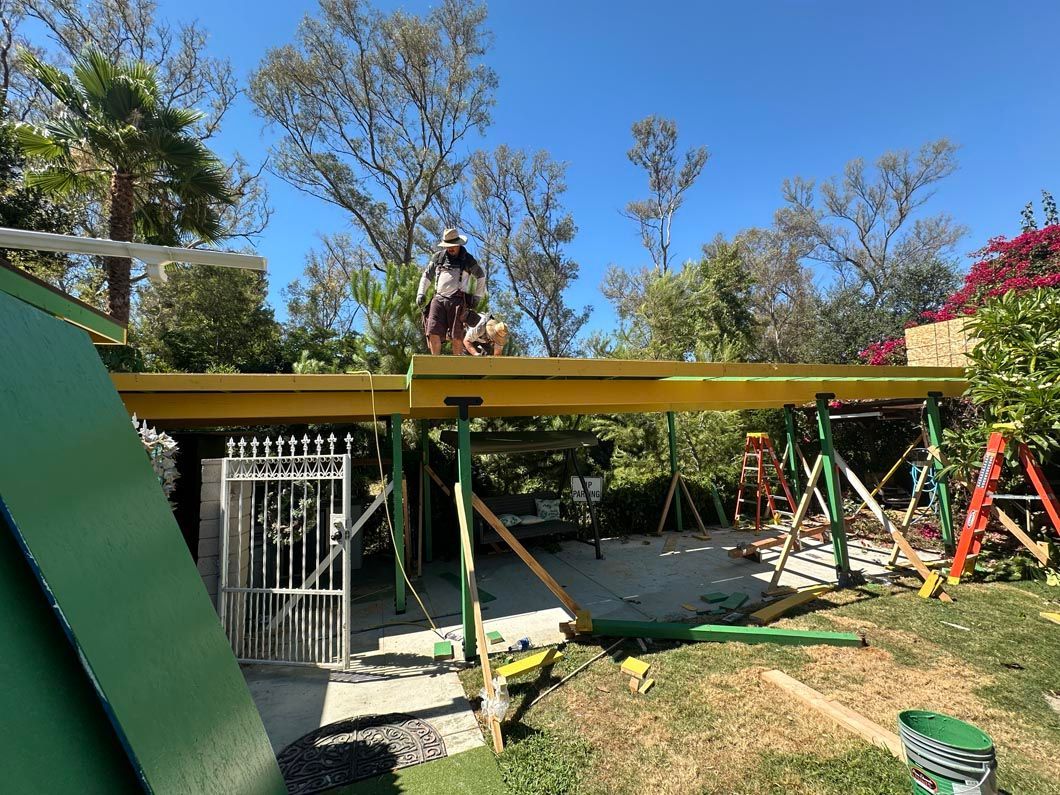 Person standing on a yellow construction beam above a backyard with trees and a metal gate