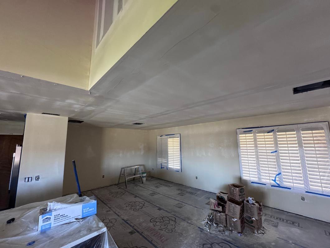 Unfinished room under renovation with gray ceiling, beige walls, windows, ladders, and construction materials scattered on floor
