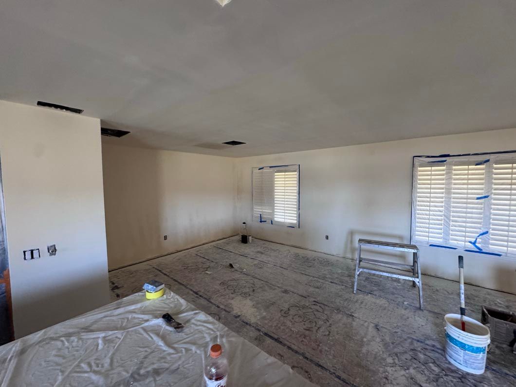 Empty room under renovation with tools, plastic sheeting, and two windows with blinds.