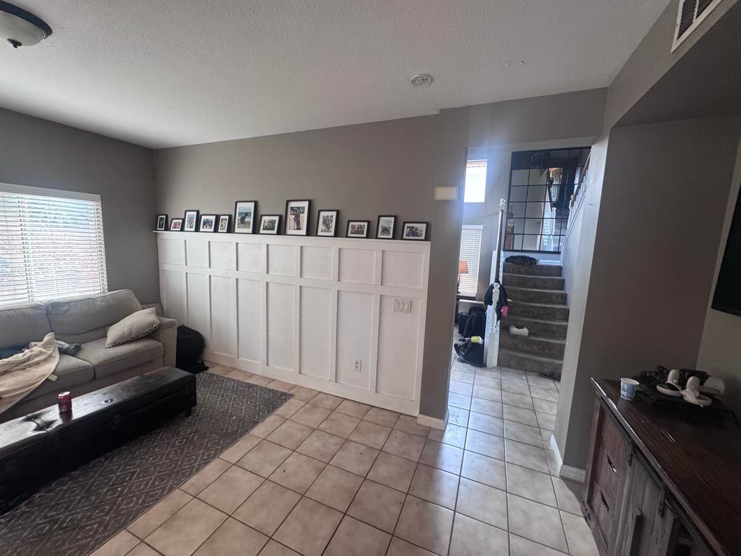 Living room with white cabinets, gray sofa, tiled floor, framed photos, and stairs in the background