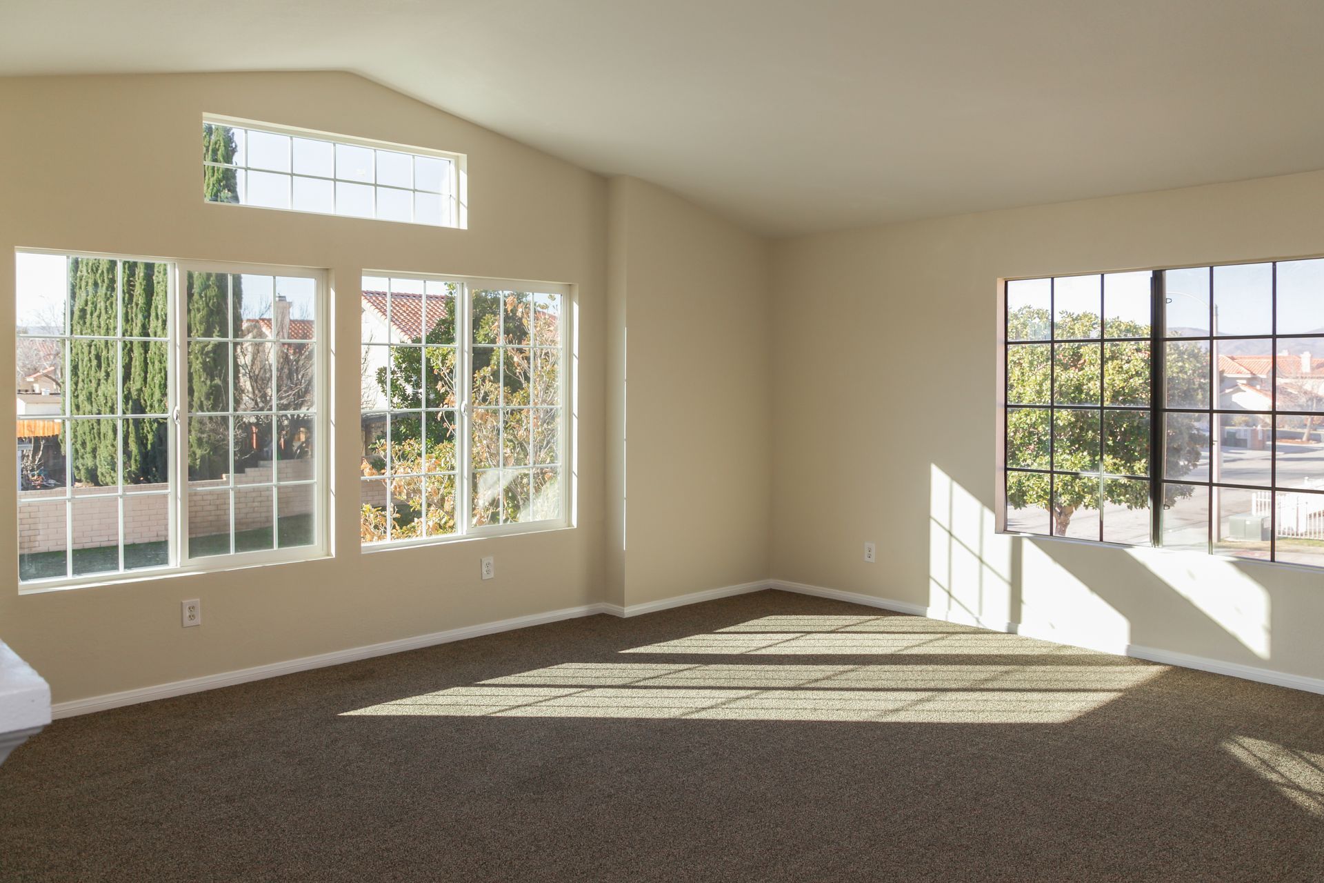 Empty beige living room with large windows, carpet, and sunlight streaming across the floor