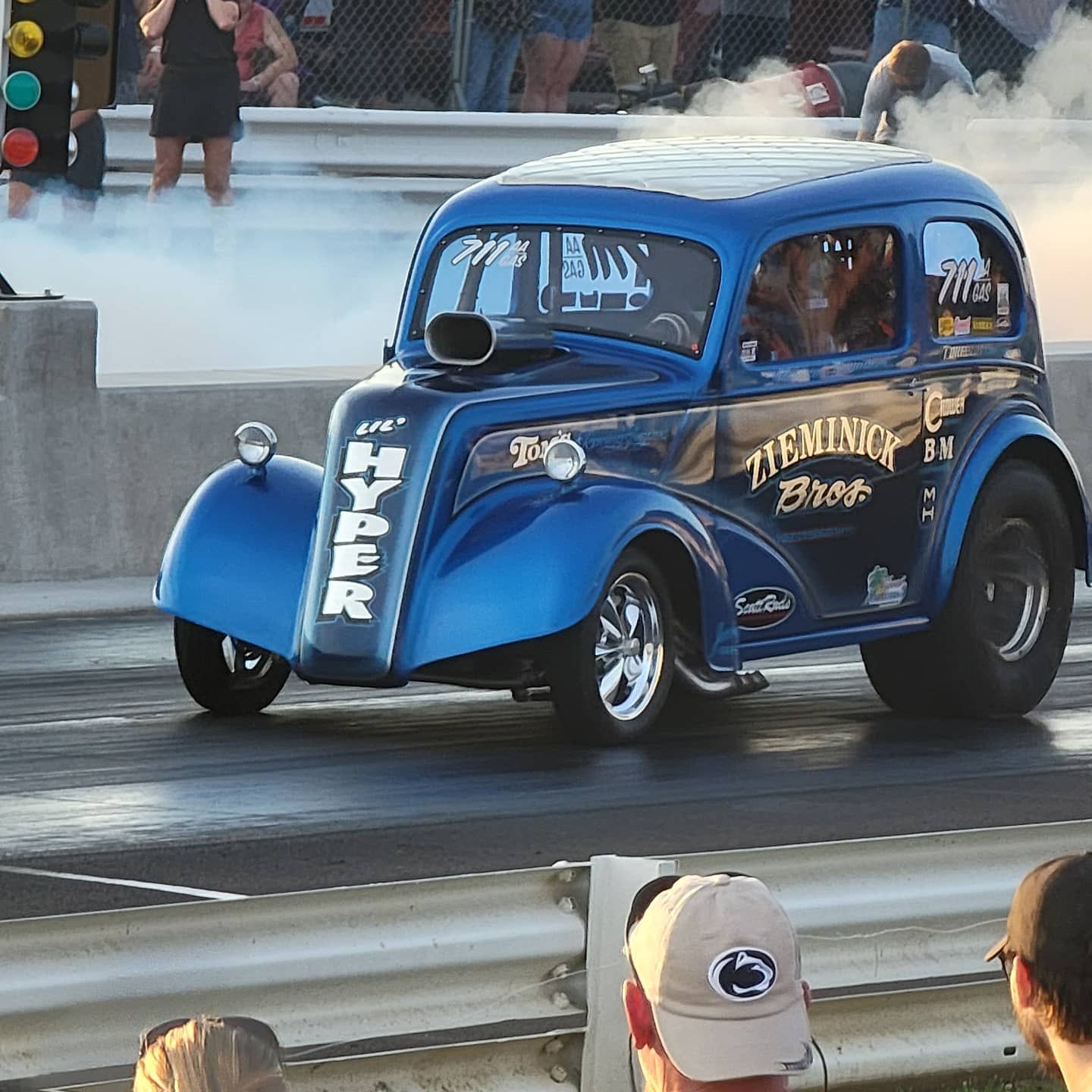 Blue vintage hot rod launching at a drag strip, smoke trailing from the rear tires.