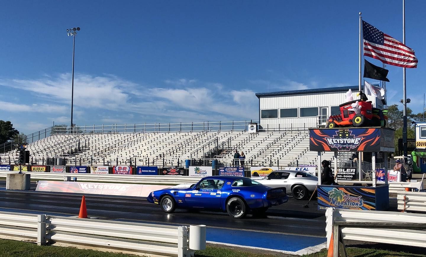 Blue race car at a drag strip with a grandstand and American flag under a clear sky.