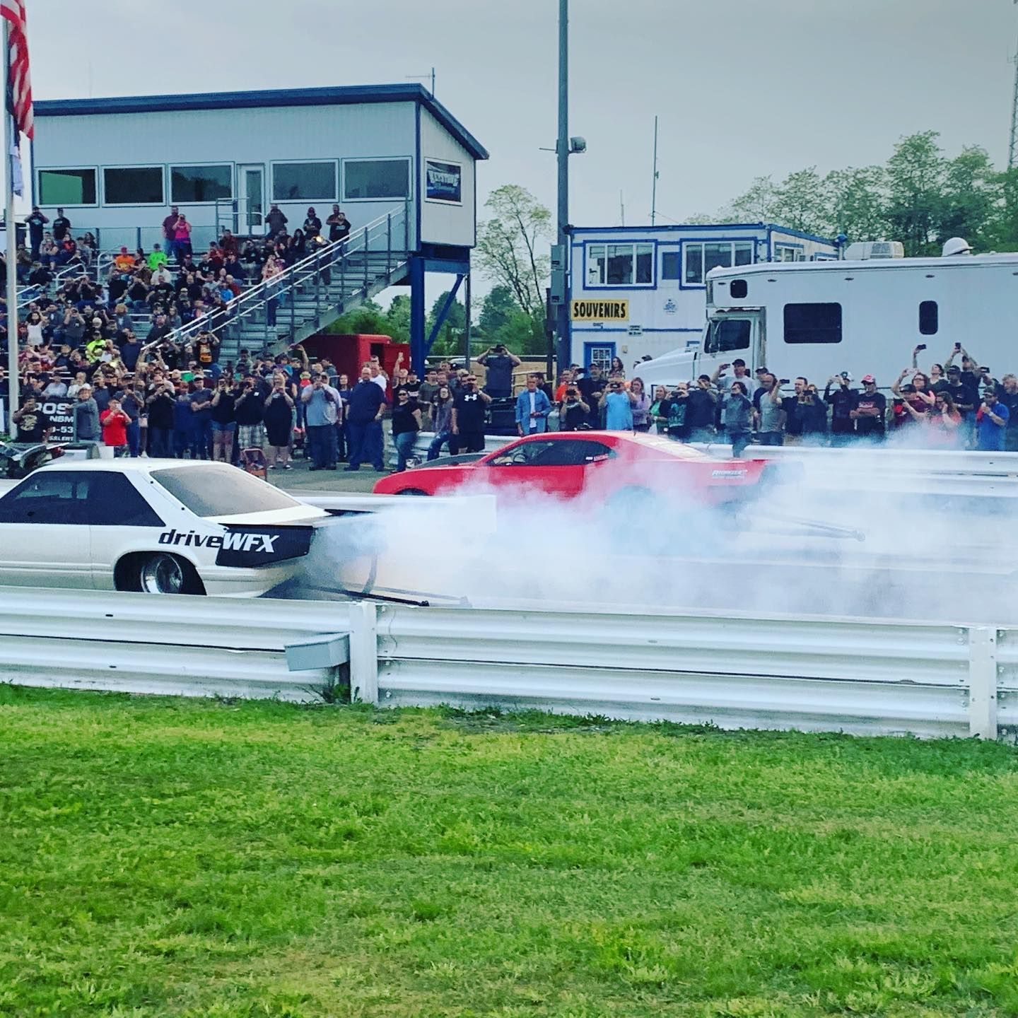 Two drag cars racing side by side, one red, with tire smoke on a track before a crowd.