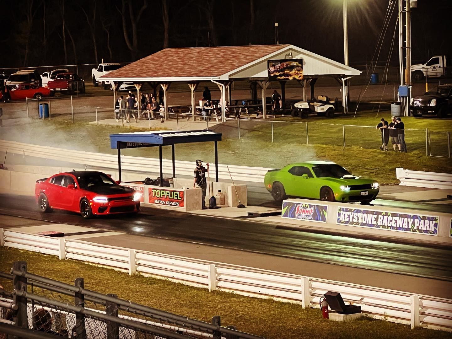 Nighttime drag strip with a red and a green muscle car staged near the start line