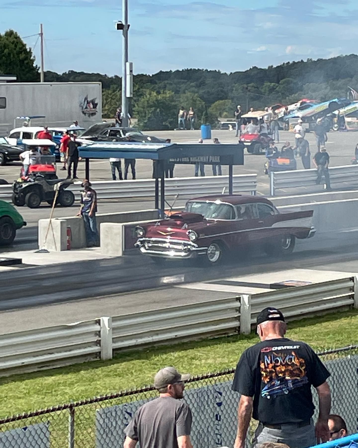Classic cars doing burnouts at a drag strip, with smoke and spectators behind a fence.