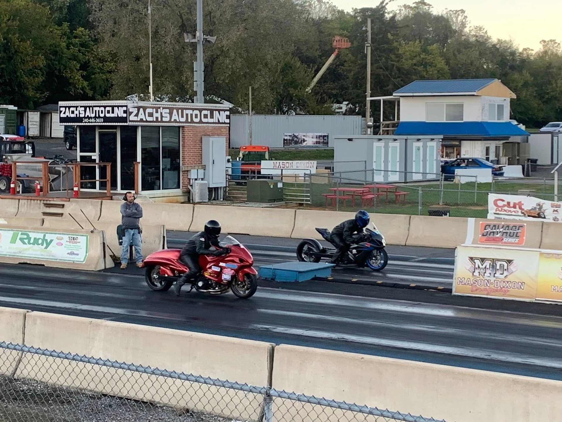 Two motorcycles racing side by side on a drag strip with a track official nearby.