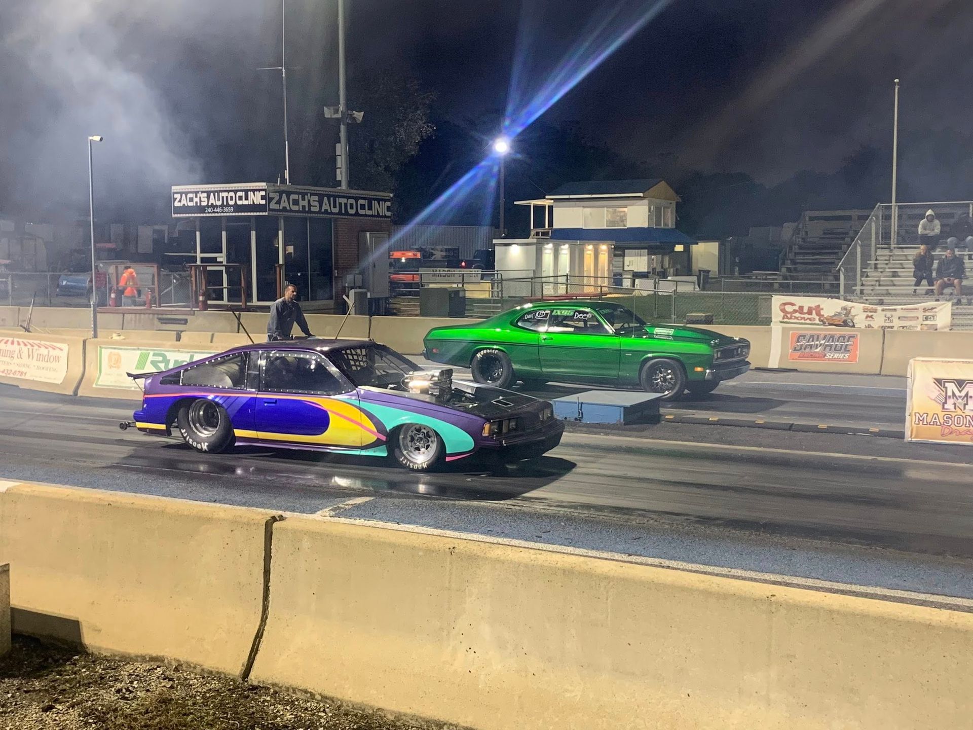 Two colorful drag racing cars line up on a nighttime track behind a concrete barrier