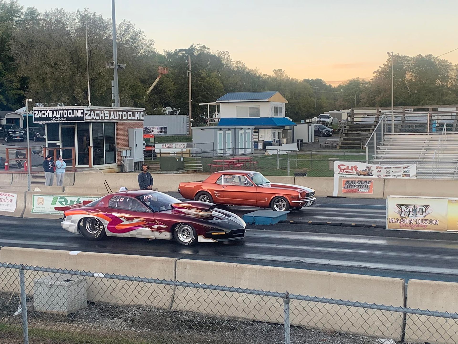 Two classic drag cars race side by side on a track at dusk.