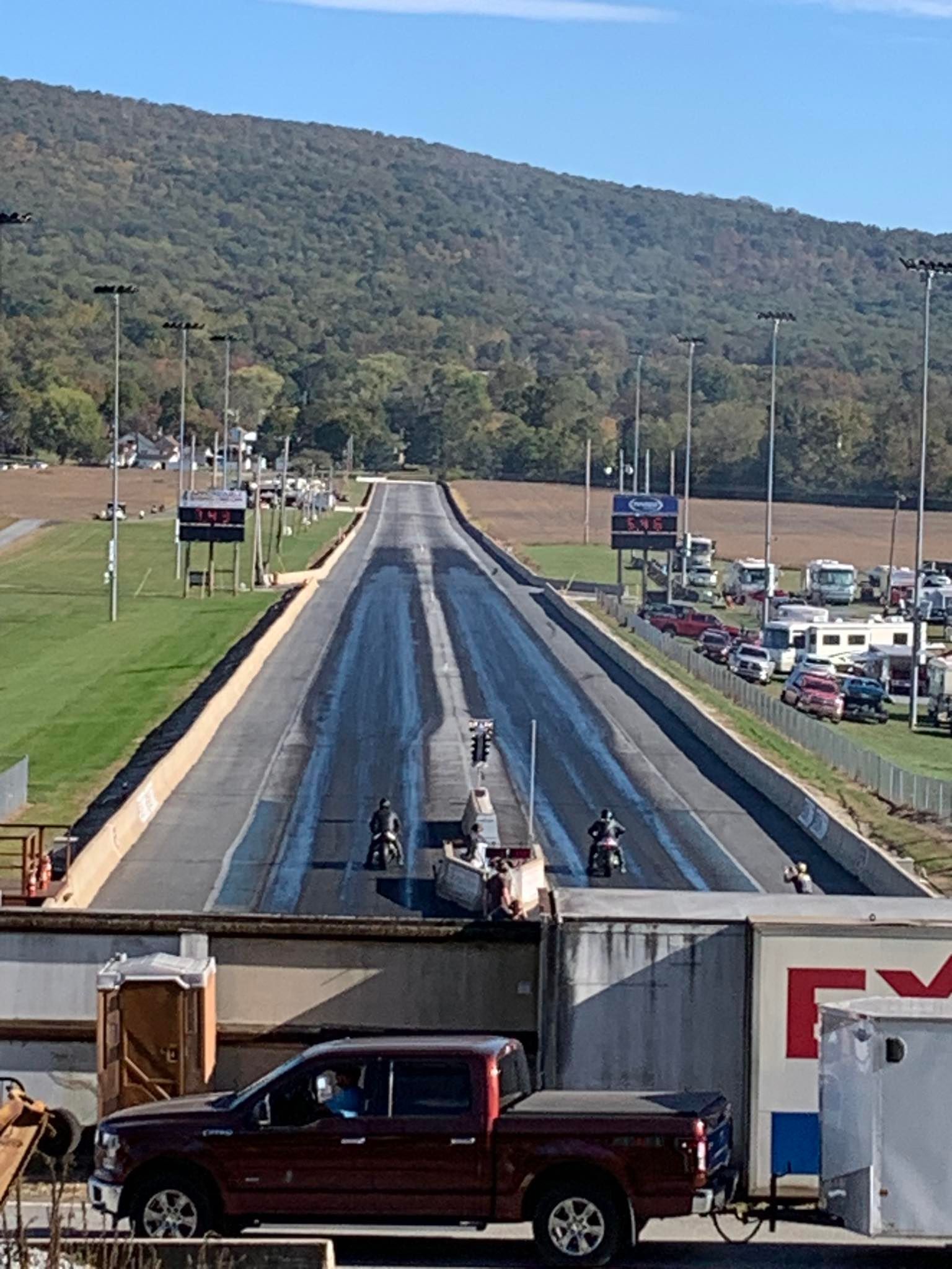 Drag racing strip with two cars staged at the start, hills and parked vehicles in the background