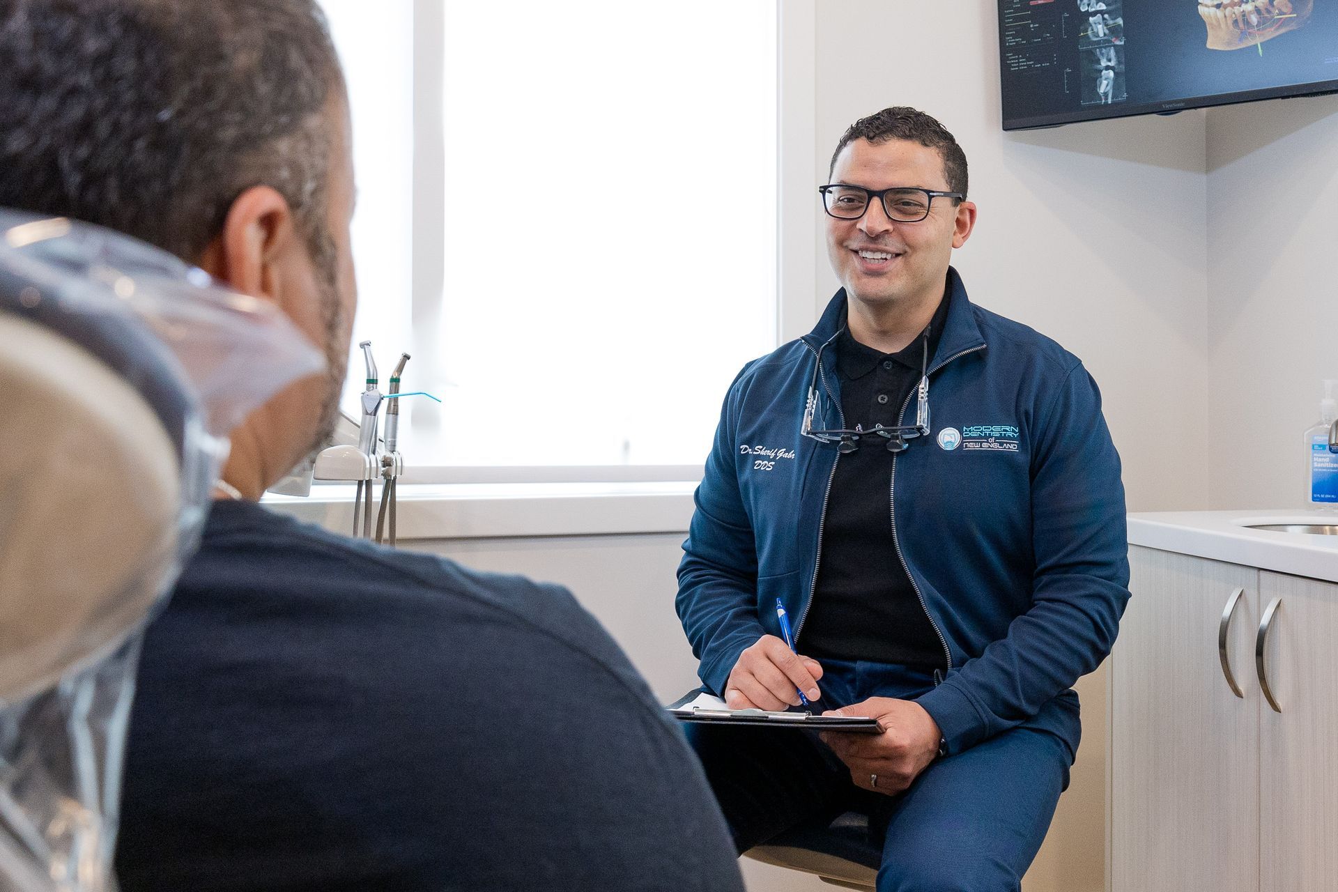 A man is sitting in a dental chair talking to another man.