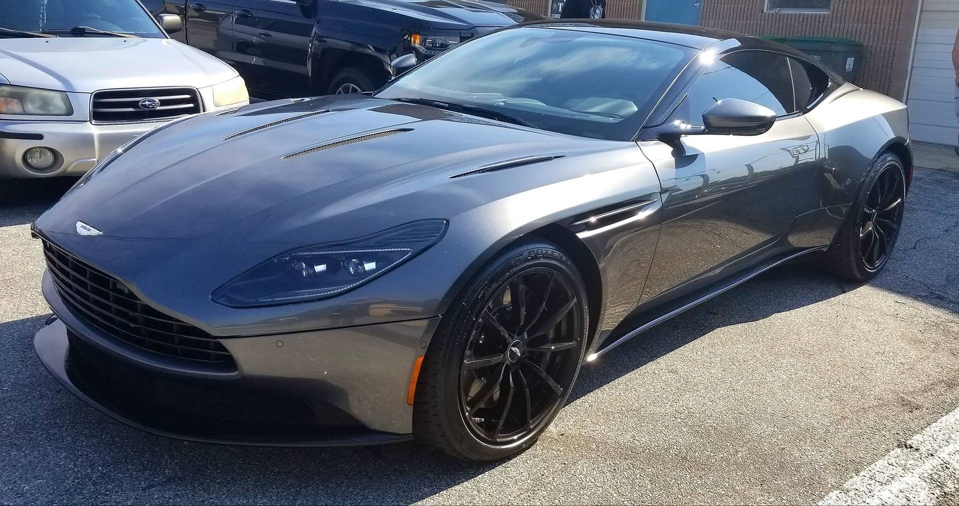 Dark gray sports car parked outdoors in sunlight, front three-quarter view.
