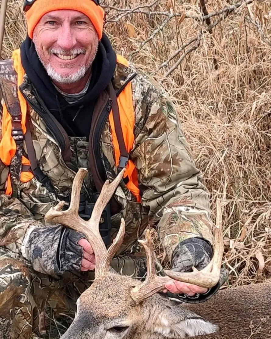 Hunter in orange gear smiles beside a deer with antlers in a brushy field.