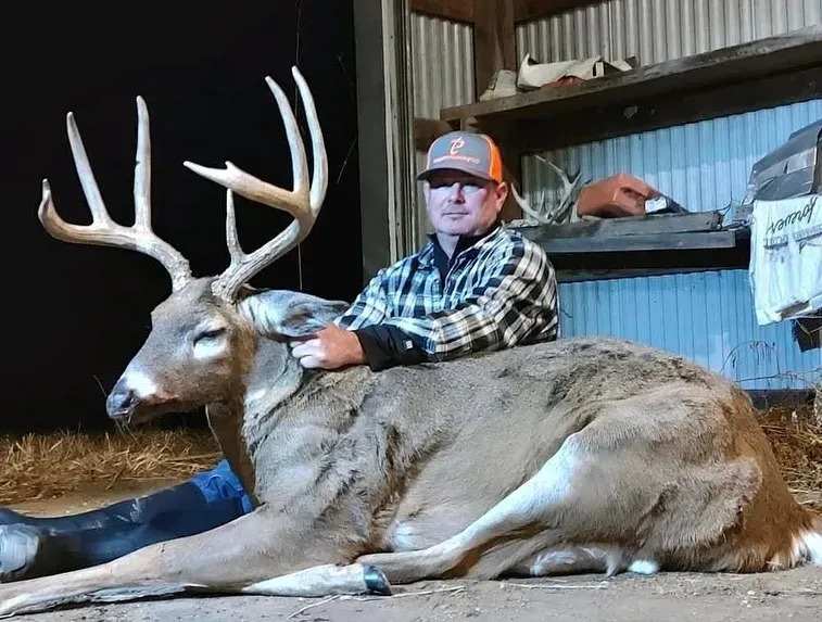 Man posing beside a large buck with antlers in a barn, warm light on both.