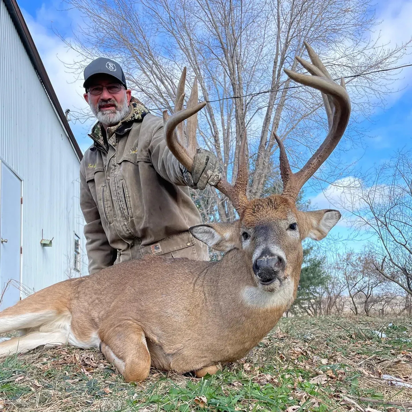 Hunter in orange gear smiles beside a deer with antlers in a brushy field.