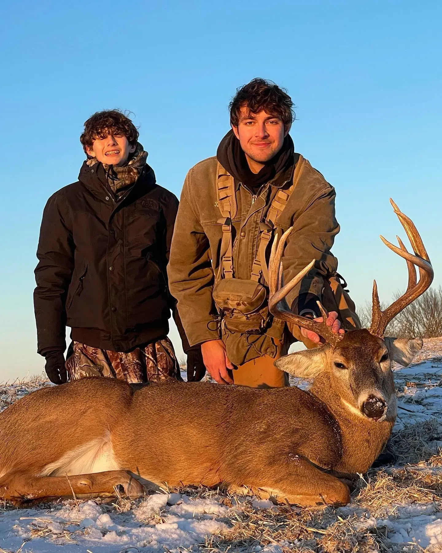 Two hunters stand behind a deer lying on snow under a clear blue sky.