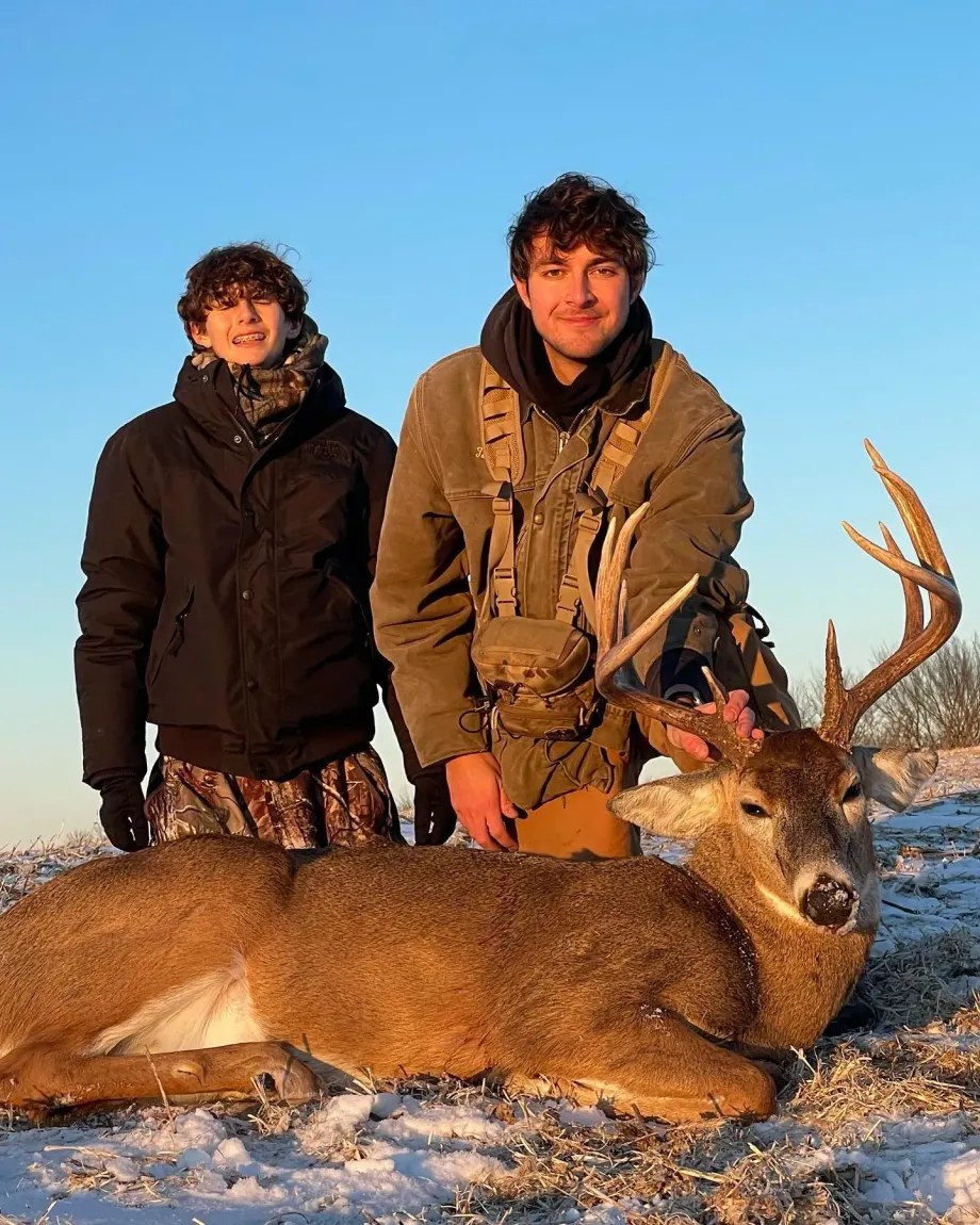 Two hunters stand behind a deer lying on snow under a clear blue sky.