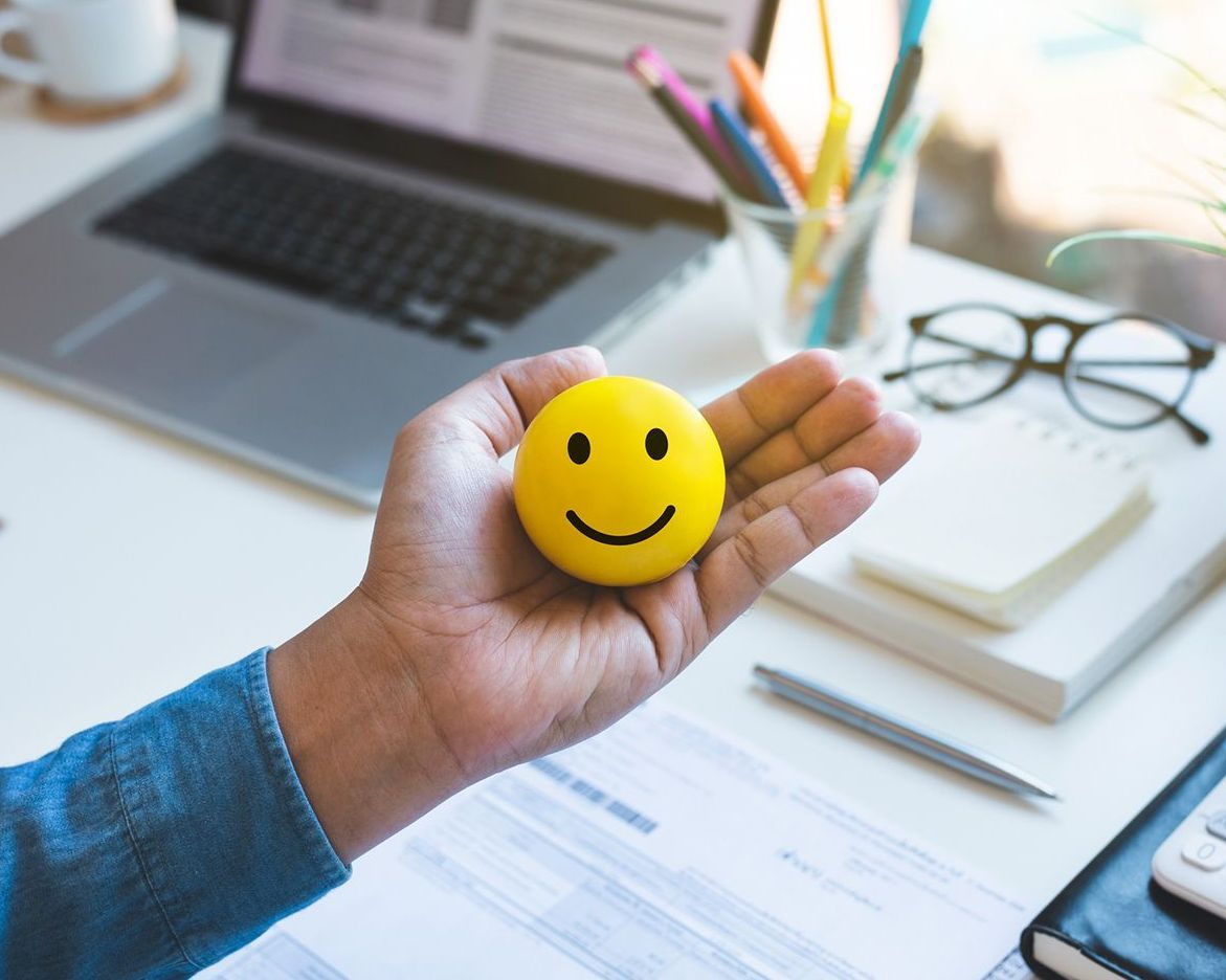 A person is holding a yellow smiley face in their hand in front of a laptop.
