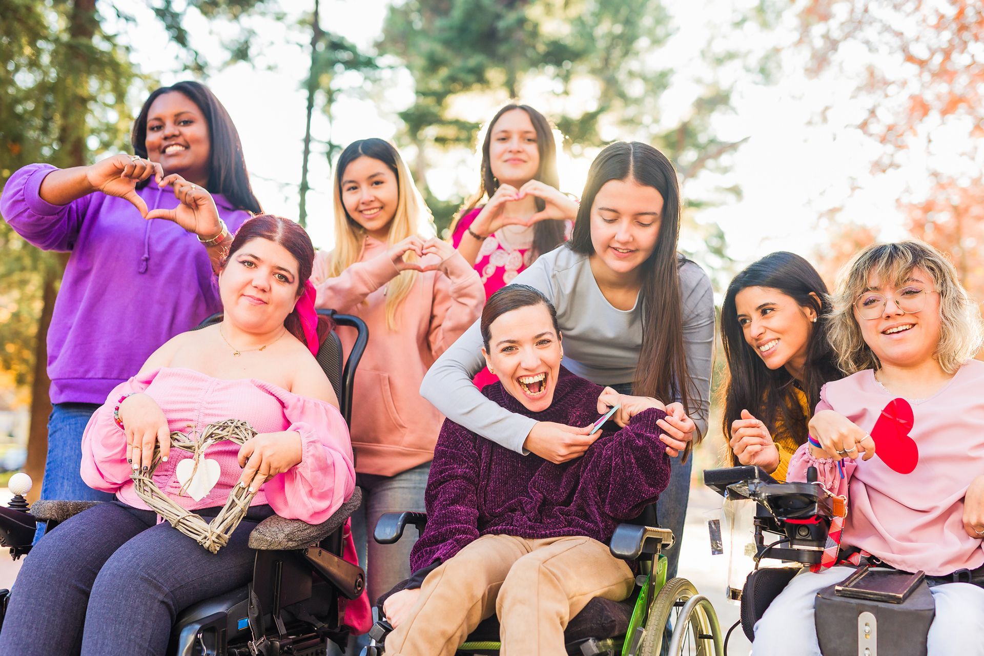 Group of smiling people in wheelchairs outdoors, posing together and making heart shapes with their hands