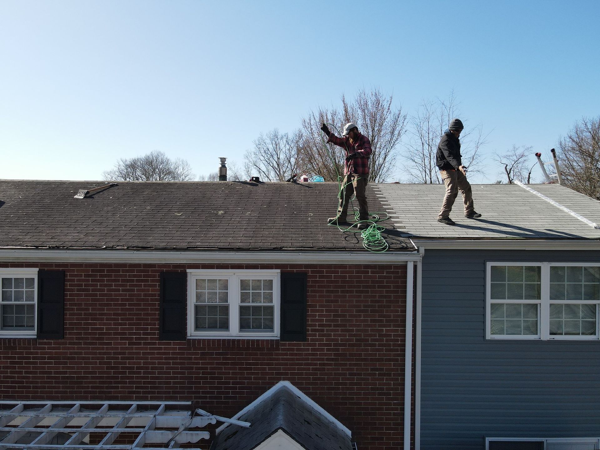 Two workers on a sloped roof, one kneeling near tools, with brick houses below and a blue sky above
