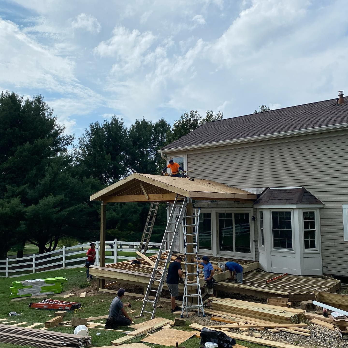 Workers installing a wooden porch roof extension on a house, with ladders and lumber in the yard.