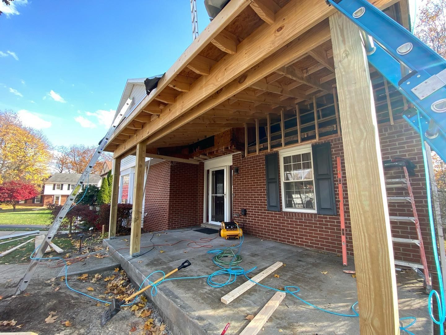 Front porch under construction with exposed roof beams, brick house wall, and ladder on the right