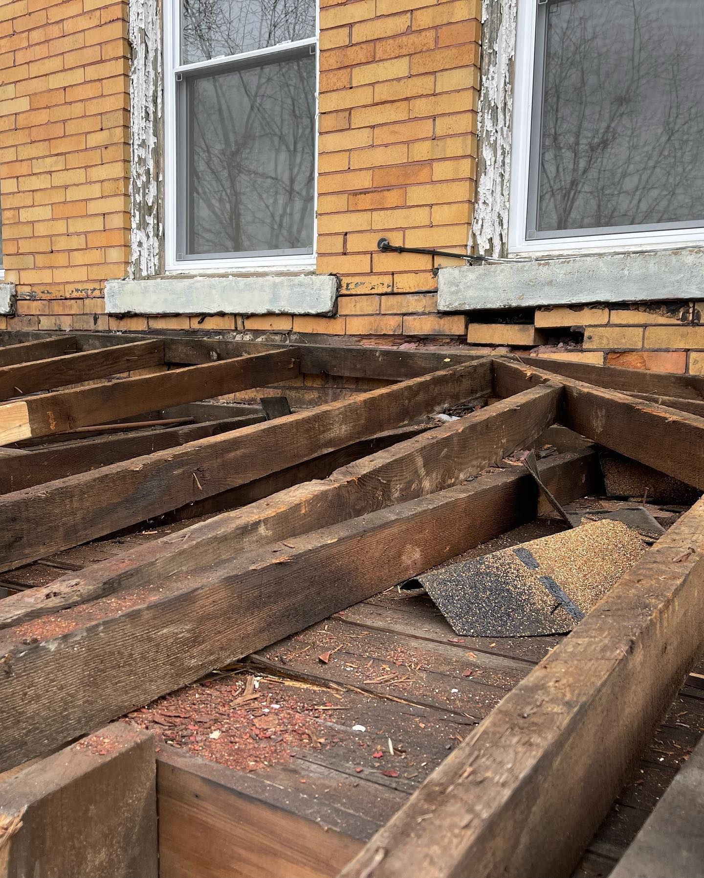 Collapsed wooden porch deck beside a brick house with two windows and scattered debris