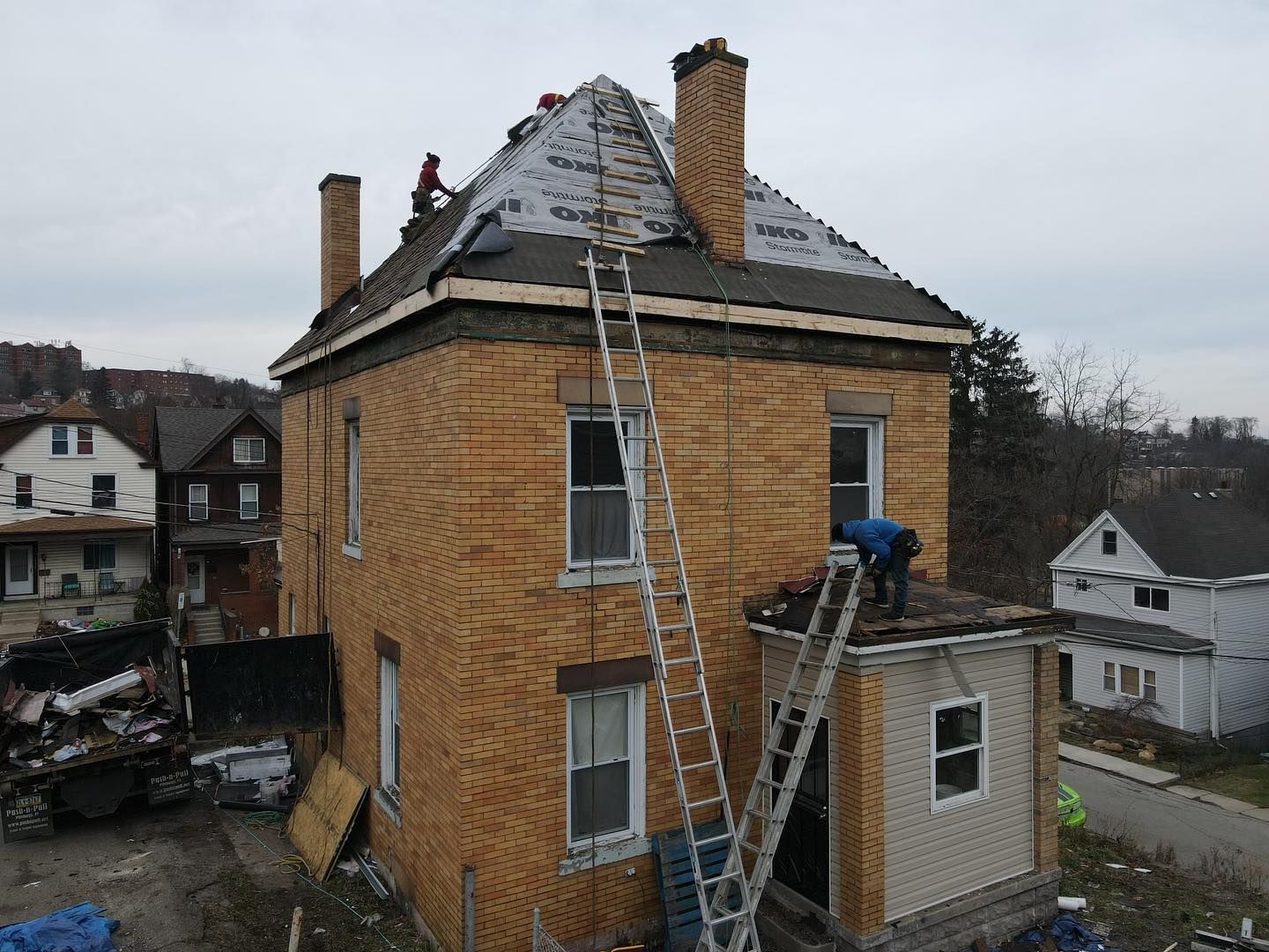 Workers on ladders repairing the roof and exterior of a brick house after damage.