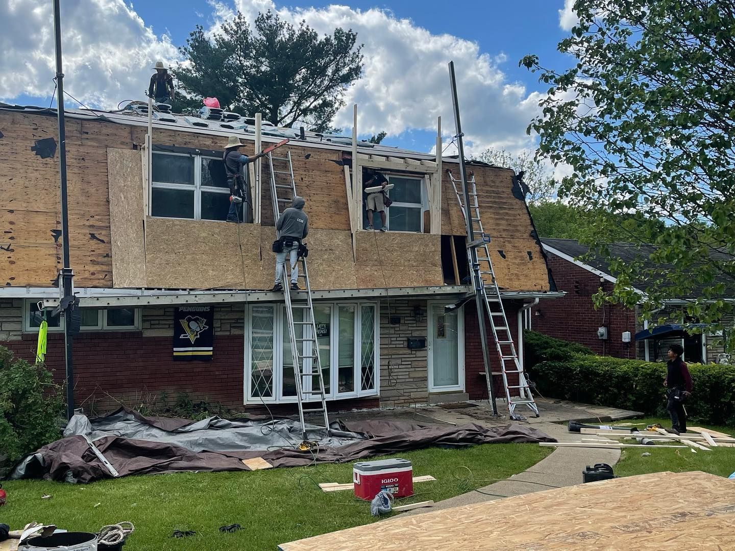 Workers on ladders repairing storm-damaged two-story brick house with shattered upper windows and debris below