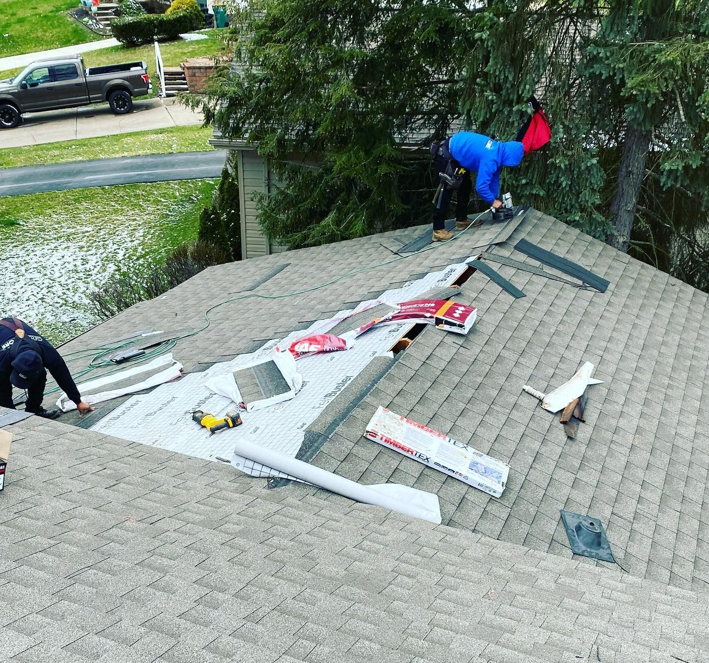 Workers repairing a shingled roof with tools and materials spread across it, trees and street in background