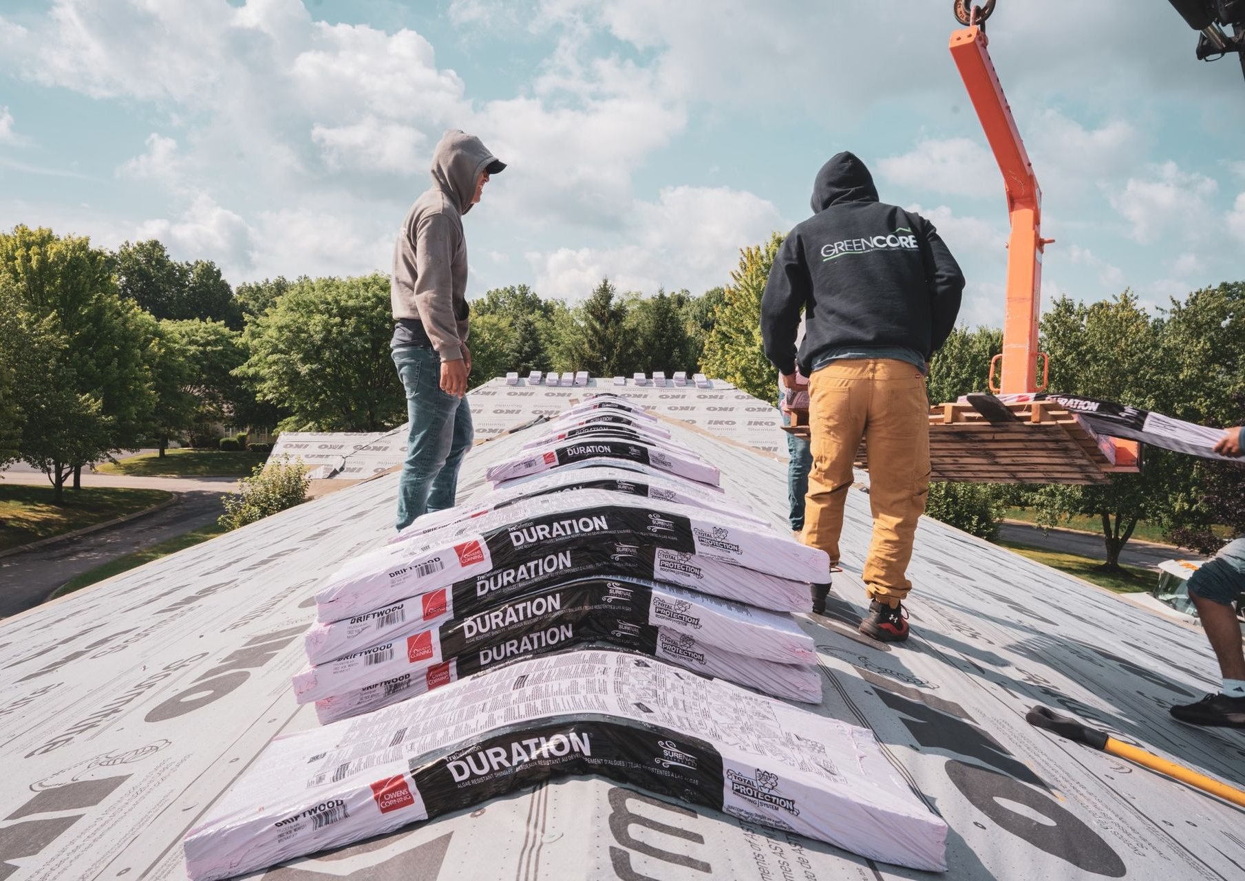 Two workers standing on a rooftop, inspecting white roofing material with an orange boom lift nearby