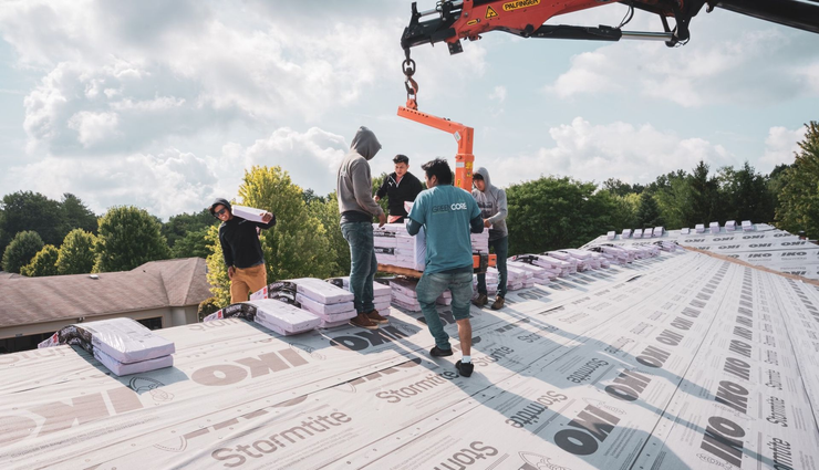 Workers on a roof guiding an orange boom lift over white roof underlayment near trees and cloudy sky