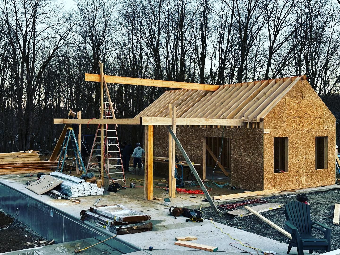 House under construction with exposed wood framing, roof trusses, and building materials scattered outside.