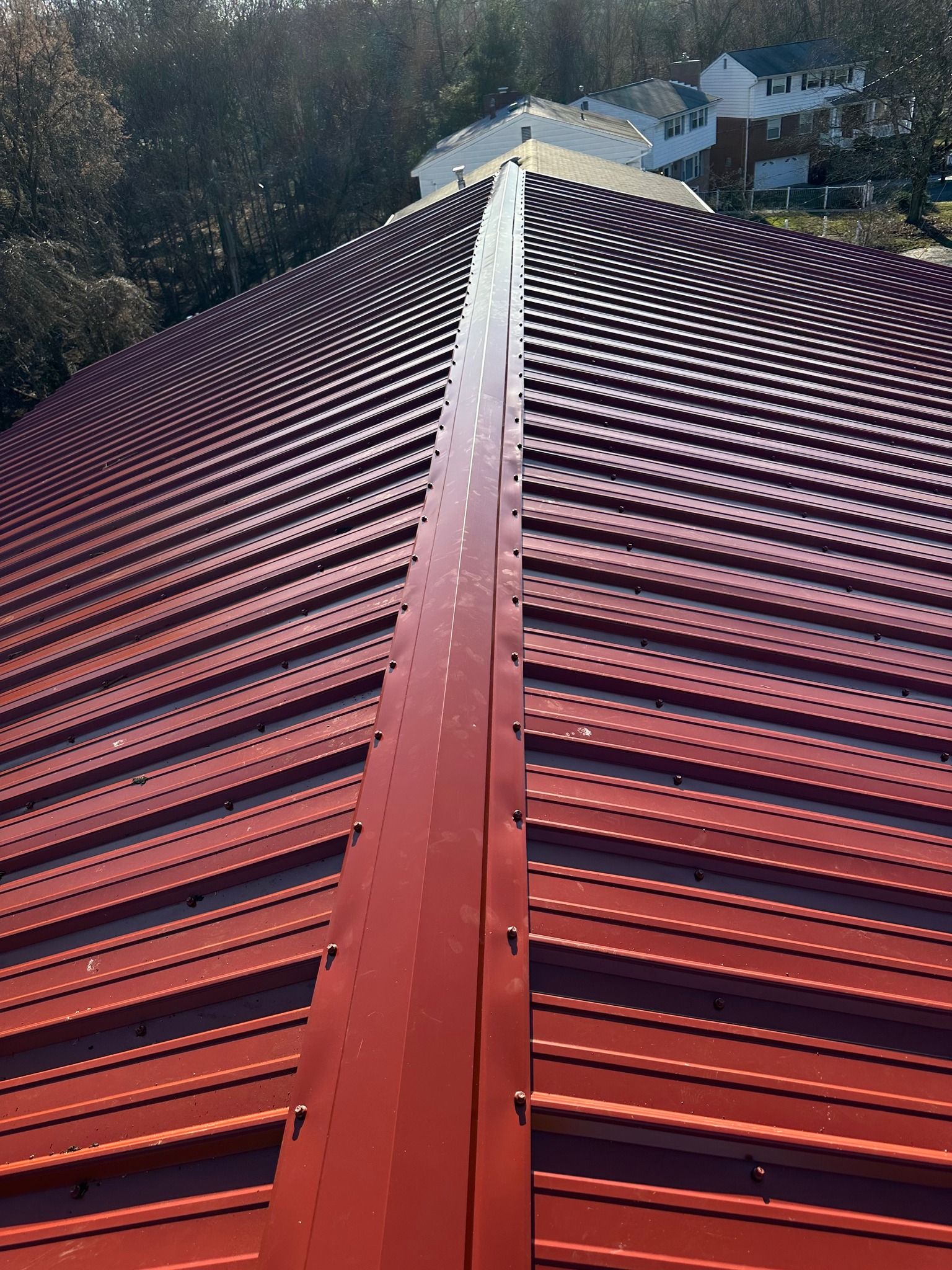 Red metal roof with a central ridge line extending toward a small house in a wooded background