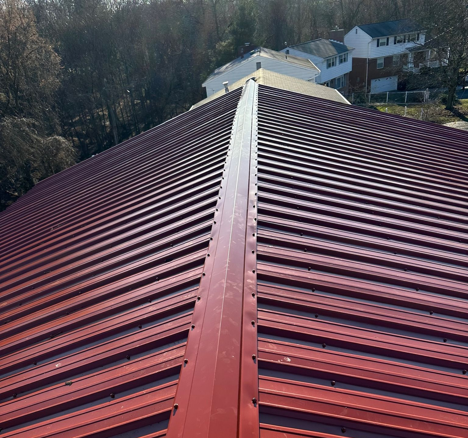 Red corrugated metal roof with a central ridge, viewed from the top toward houses and trees beyond