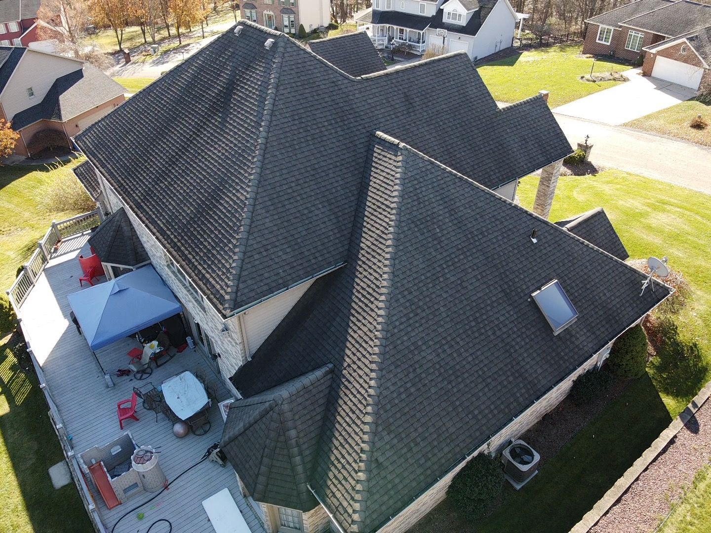 Aerial view of a dark-roofed house with a patio, lawn, and neighboring homes around it.