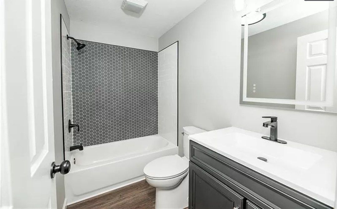 Bright bathroom with white tub and toilet, gray patterned shower curtain, and vanity with mirror.