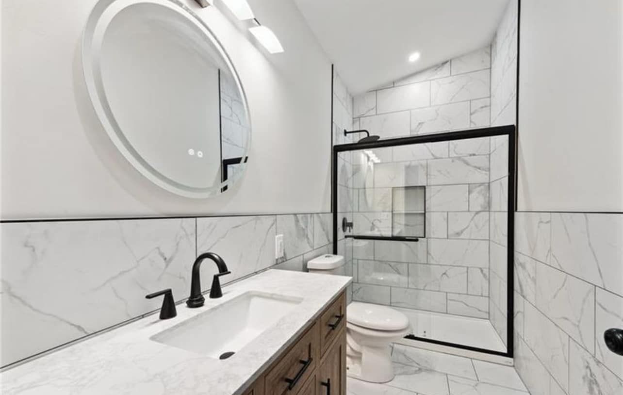 Modern white bathroom with black fixtures, round mirror, marble tile, and glass shower enclosure