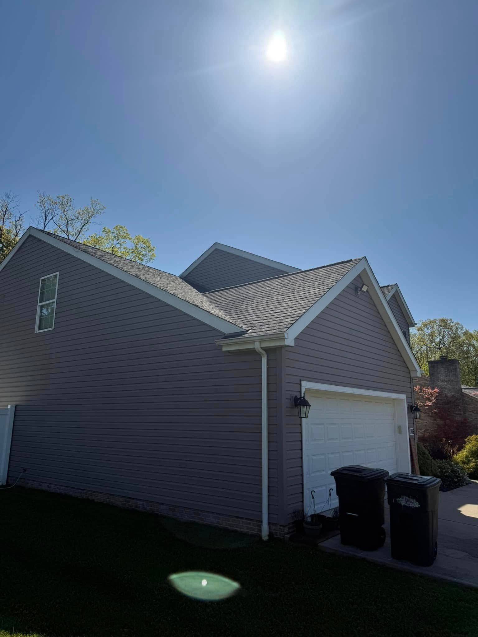 Suburban house with white garage door under bright sun, with trash bins by the driveway.