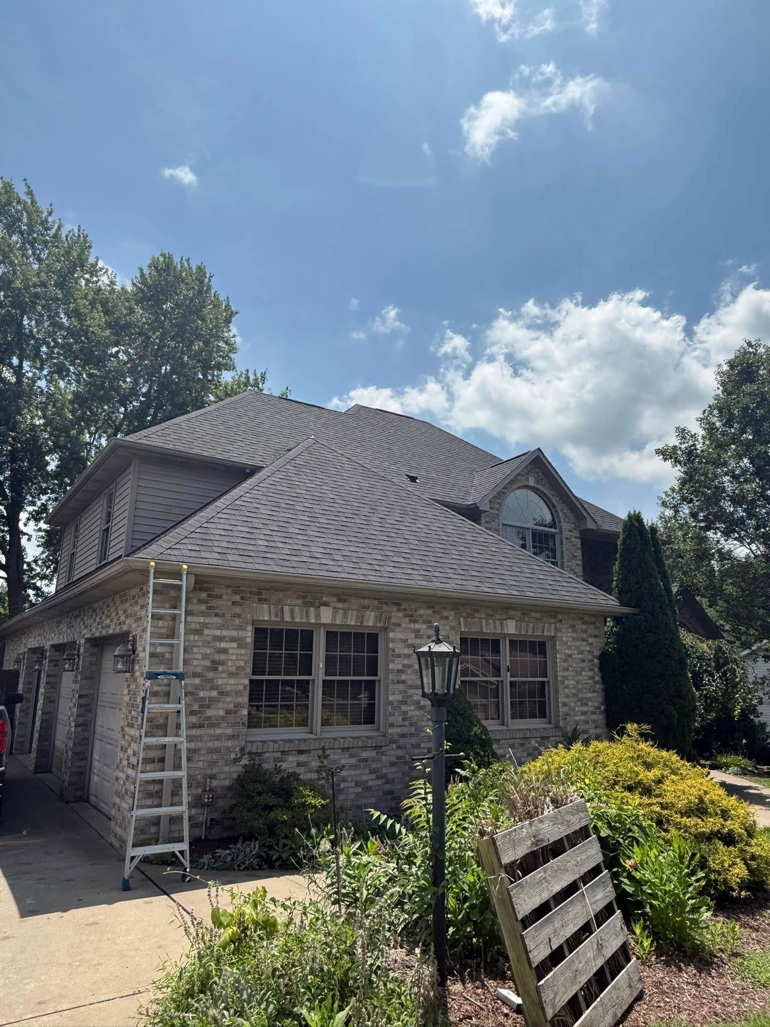 Stone house with a wavy gray roof, ladders by the wall, and a sunny garden under a blue sky