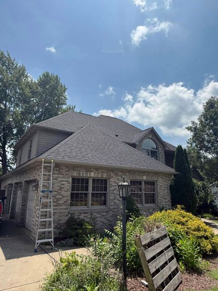 Stone house with a wavy gray roof, ladders by the wall, and a sunny garden under a blue sky