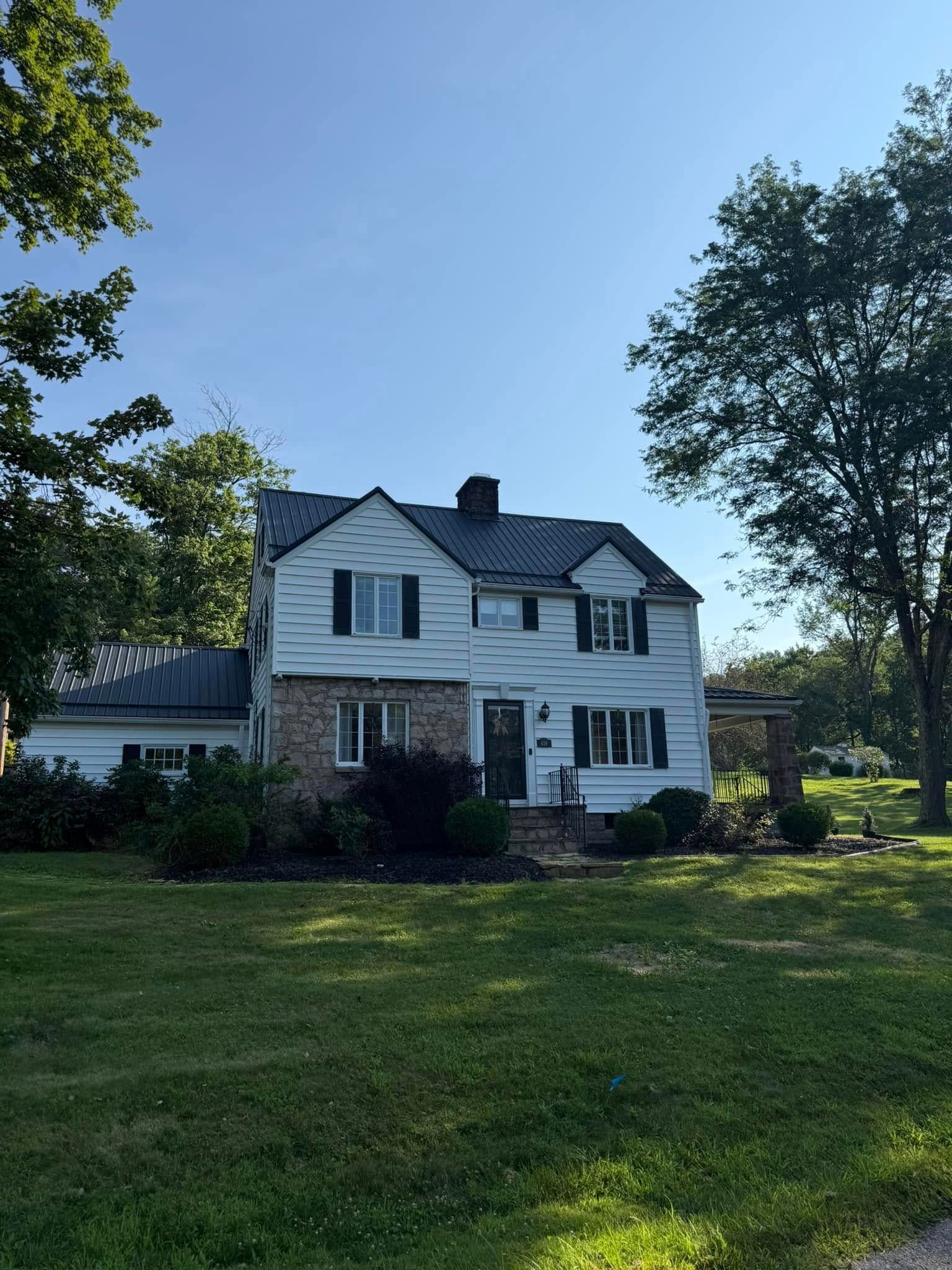 White two-story house with dark roof and chimney, surrounded by trees and a green lawn under a clear blue sky