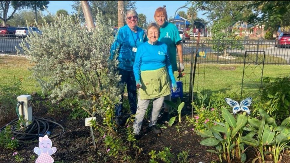 Three people in teal shirts and aprons stand in a garden.