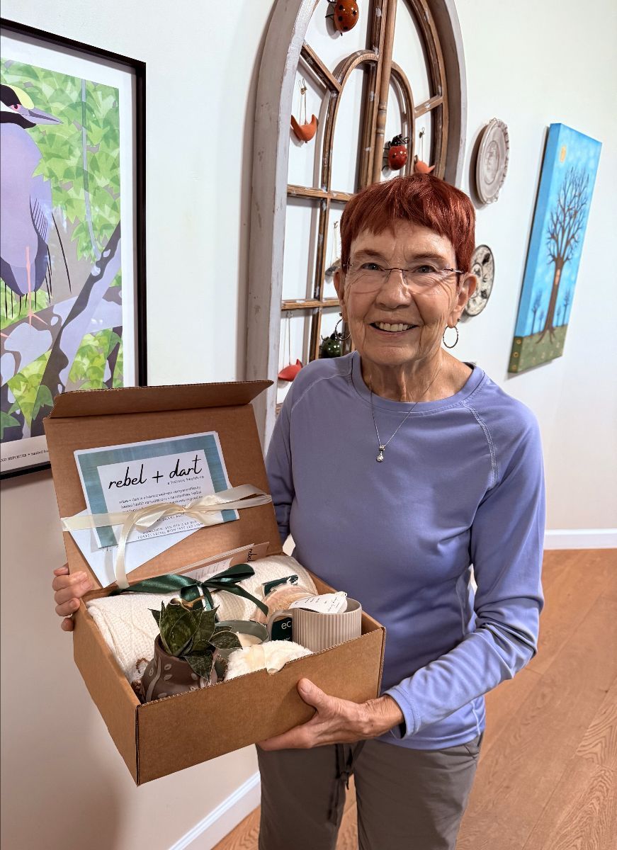 Woman holding gift box, smiling. Interior setting with artwork and window. Box contains items wrapped in ribbon and a plant.