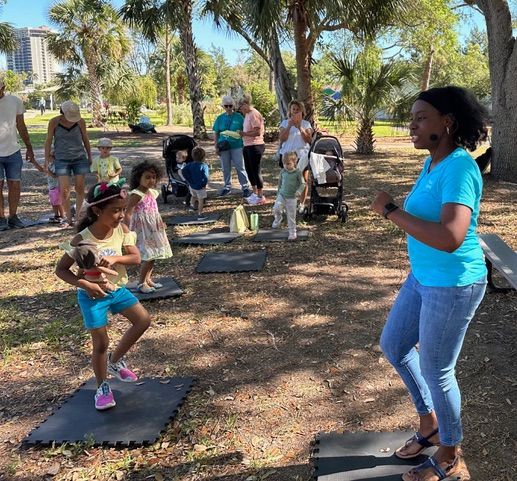 A woman in a blue shirt is standing in front of a group of children in a park.